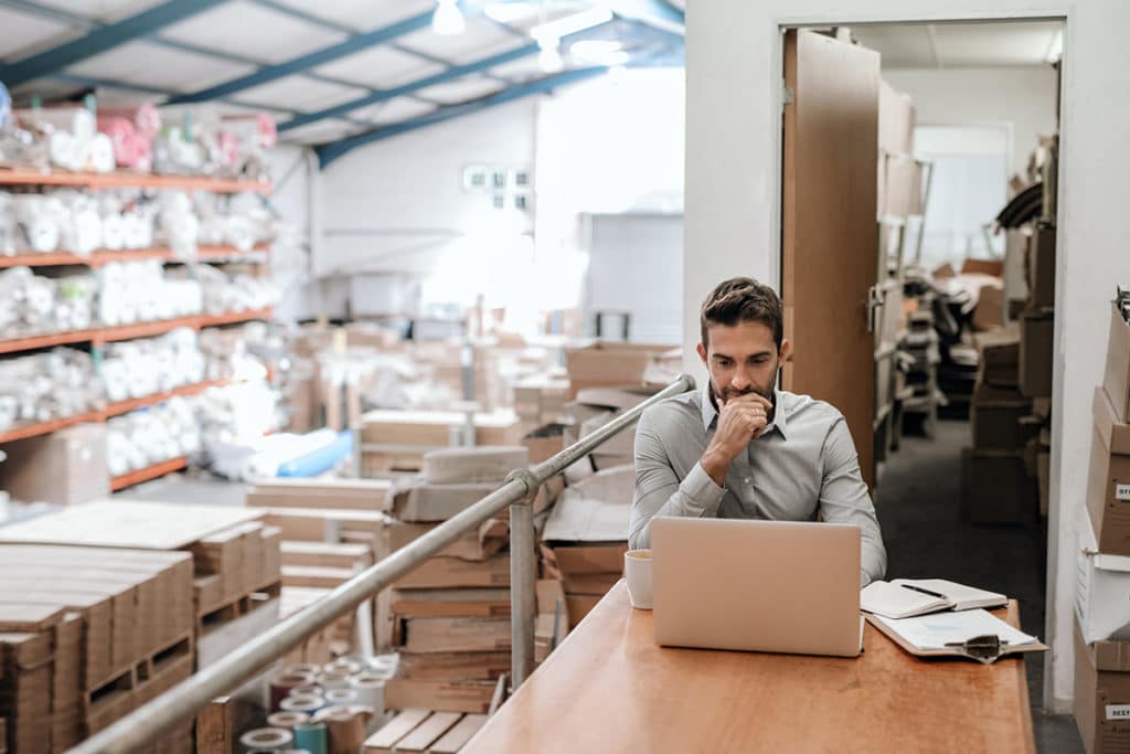 un professionnel est assis sur un bureau avec son ordinateur portable, un café et des carnets. il surplombe un entrepôt remplis de marchandises.