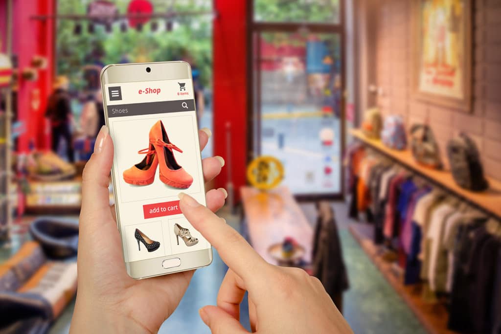 dans un magasin, une femme tient un téléphone portable sur lequel il y a à l'écran un e-Shop avec une paire de chaussures à talons oranges ainsi que le bouton "add to cart" sur lequel elle est en train de mettre son doigt.