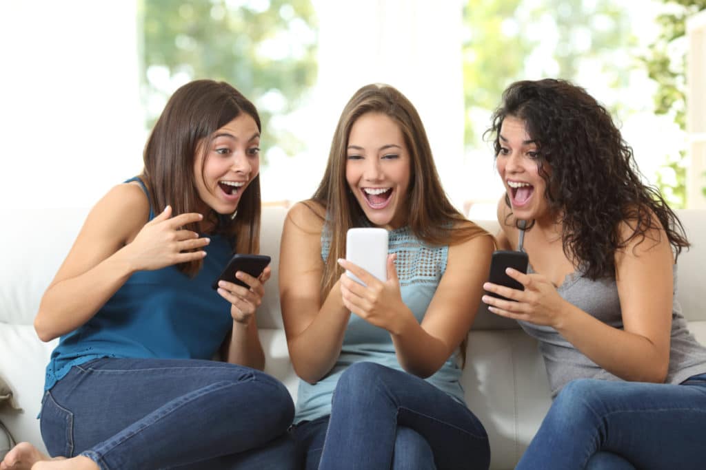 3 jeunes femmes assises sur un canapé, regarde leur téléphone portable et souriant la bouche grande ouverte.