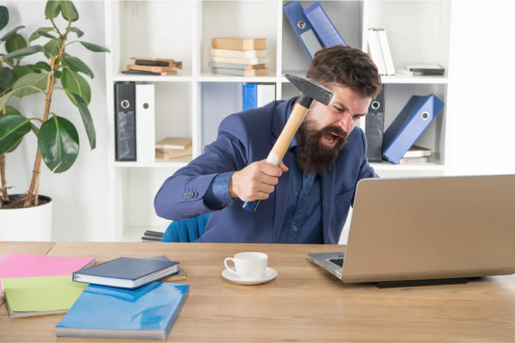 un homme tient un marteau à la main assis à son bureau. il est sur le point de taper son ordinateur portable avec en faisant une grimace de colère.