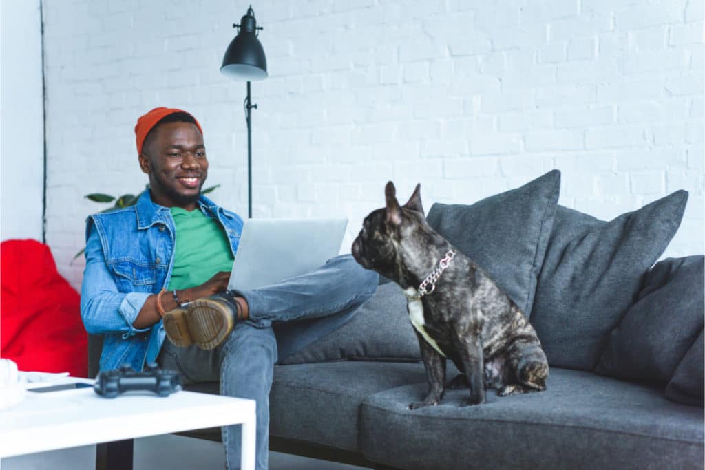 un homme de couleur noir est posé sur son canapé avec son ordinateur portable sur ses jambes et sourit à son chien assis lui aussi sur le canapé gris. il a une jambe repliée et posée sur l'autre. il porte une veste en jean et un bonnet orange.