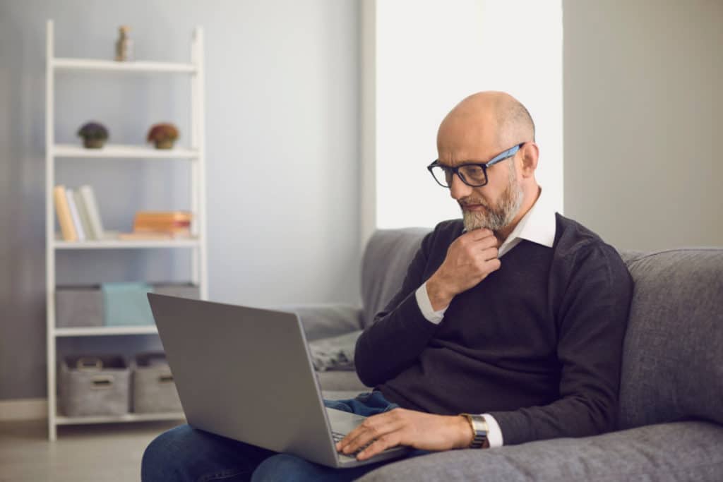 un homme âgé avec des lunettes noires et une barbe grise est assis sur un canapé gris avec un ordinateur portable gris sur ses genoux. il se touche la barbe d'une main tandis que l'autre main est posée sur le clavier.