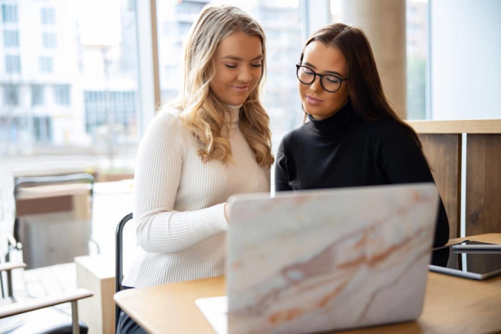 2 jeunes femmes sont côté à côte et regardent quelque chose ensemble derrière un ordinateur portable posé à un bureau.