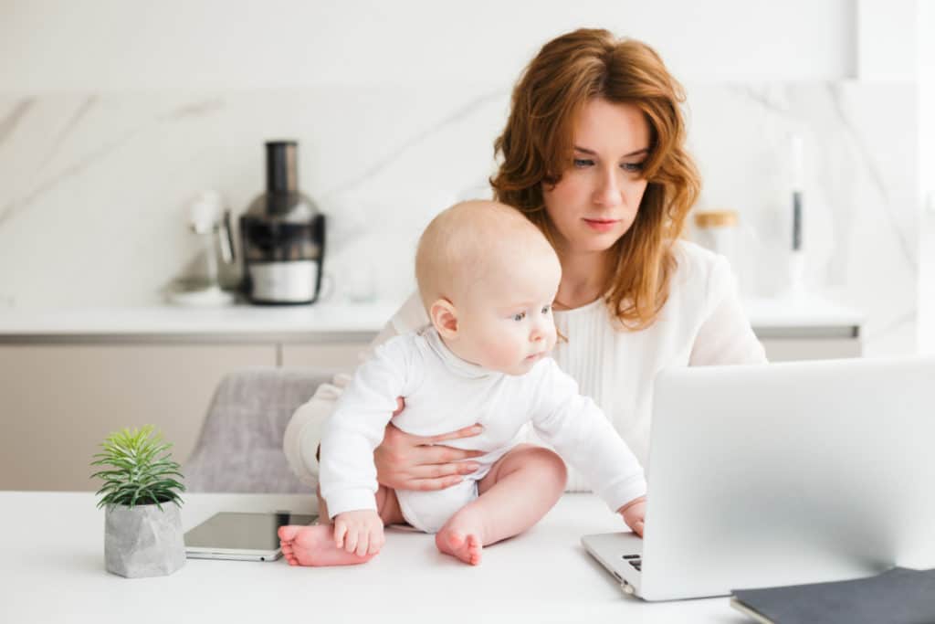 une femme rousse est assise à un table devant un ordinateur portable. elle tient un bébé en body blanc posé sur le table à côté de l'ordinateur qui regarde l'écran.