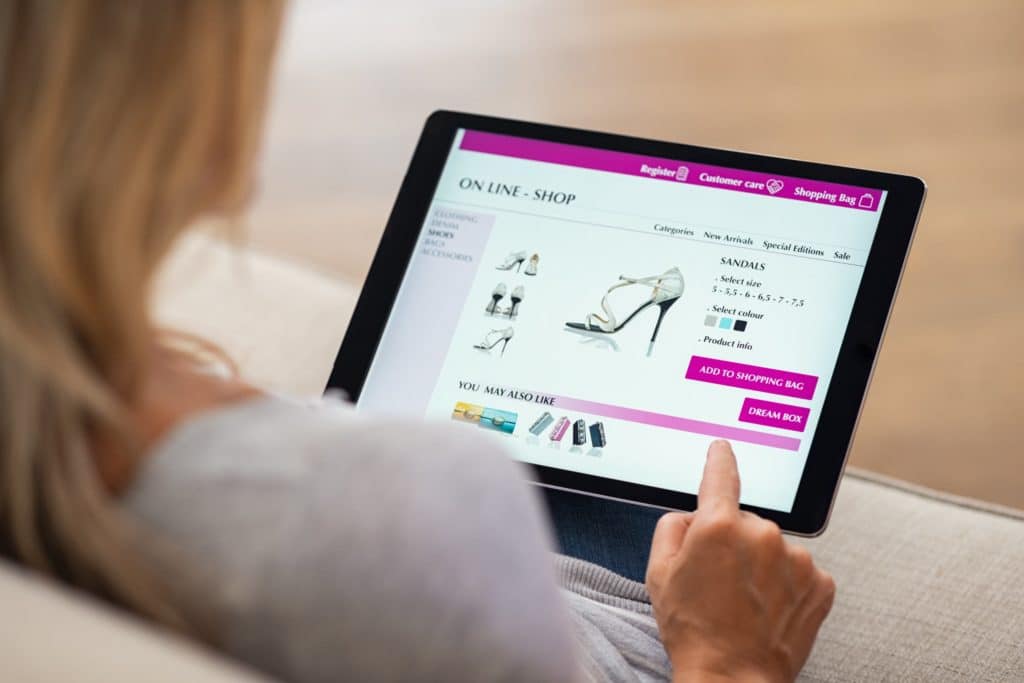 un femme a le dos posé sur une tablette sur laquelle est affiché un site e-commerce. elle est sur une fiche produit de sandales à talons.