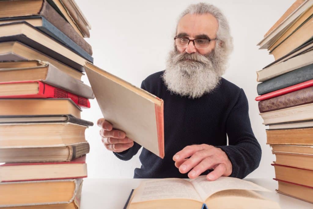 un vieil barbu avec des lunettes de vue consulte des livres anciens sur un bureau. il est entouré de piles de livres de chaque côté.