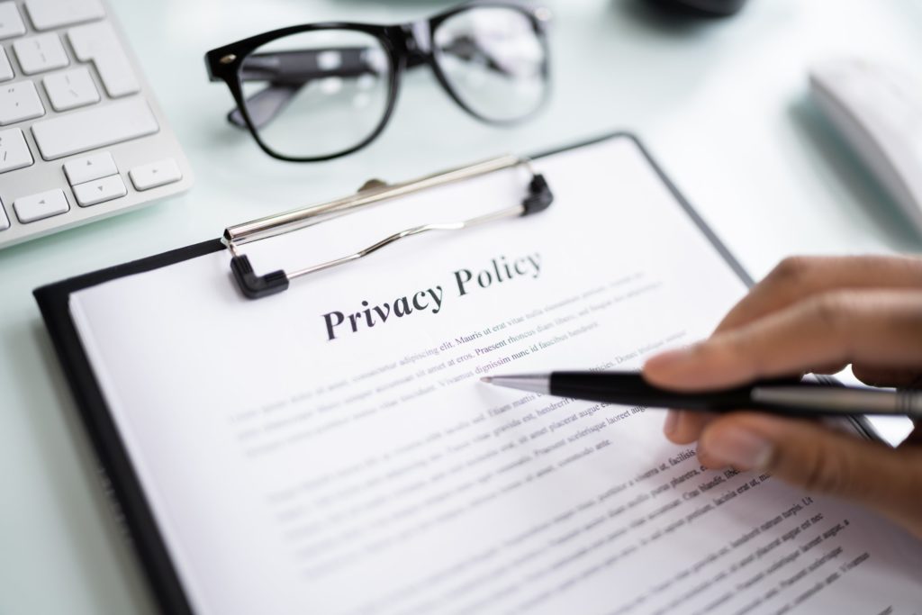 sur une table, une main tient un stylo posé sur une feuille sur laquelle figure une politique de confidentialité écrit en anglais "privacy policy". une paire de lunettes de vue noire est posée à côté ainsi qu'un clavier blanc.