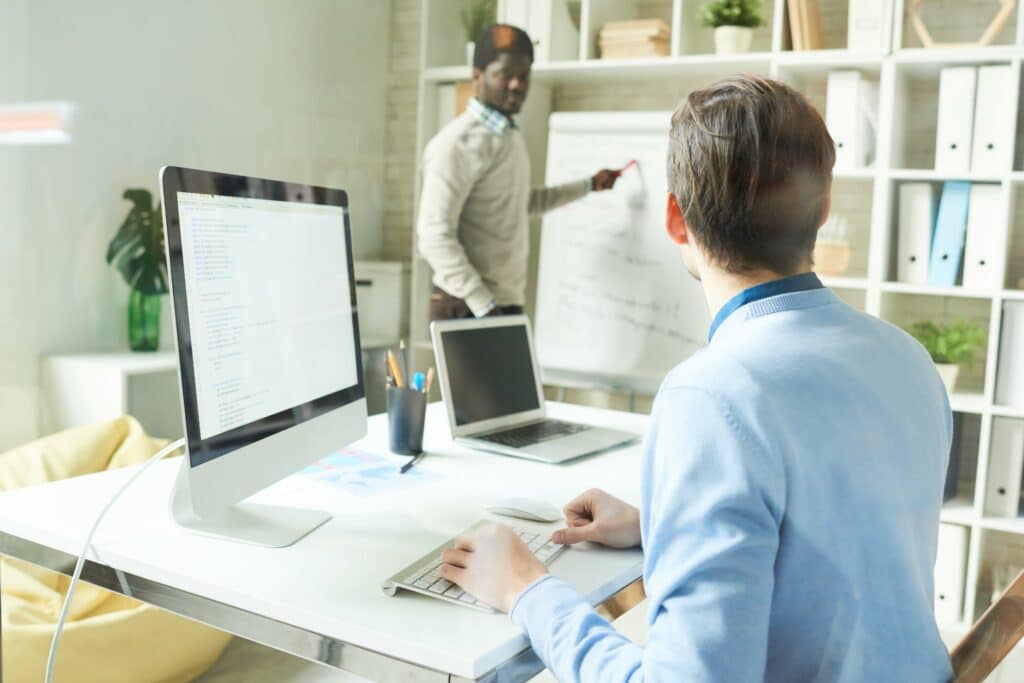 un homme en pull beige qui explique quelque chose à son collègue en chemise bleue assis devant un ordinateur.