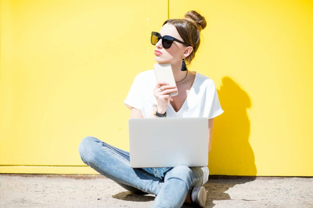 Une femme portant des lunettes de soleil assise à même le sol, adossée à un mur de couleur jaune réfléchissant devant un ordinateur portable de couleur blanche et ayant dans sa main un Smartphone de couleur blanche.