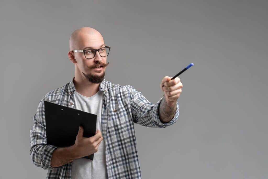 Un homme en chemise à carreaux et T-shirt , portant des lunettes tient un dossier dans une main et un stylo dans l'autre.. L'homme est entrain de montrer quelque chose.