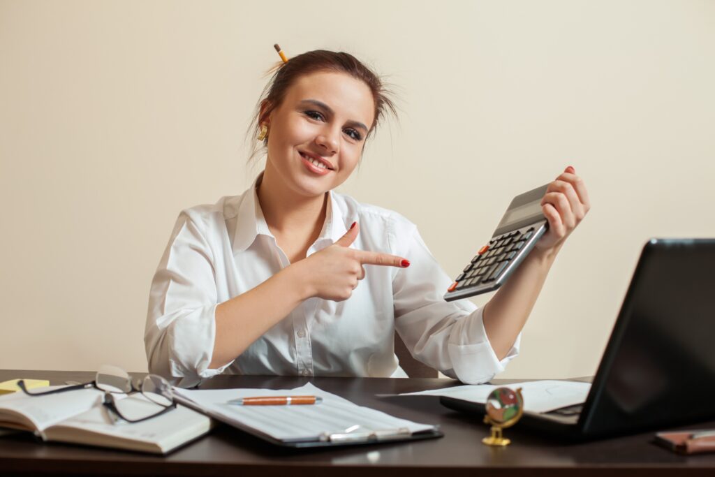 Une jeune femme souriante, pointant de son index une calculatrice qu'elle tient dans une main. Elle est assise devant un bureau sur lequel sont posés un ordinateur portable, un carnet, des lunettes, un stylo et un document.