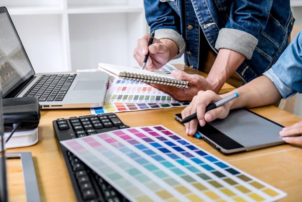 Les mains de deux graphistes travaillant ensemble sur un bureau où sont posés un ordinateur portable, des palettes de couleur, un clavier et une tablette.