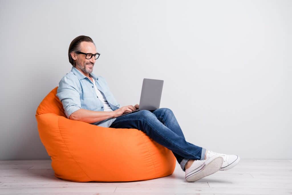Un homme d'âge mûr en tenue décontractée assis sur un fauteuil haricot de couleur orange avec son ordinateur portable sur les genoux.