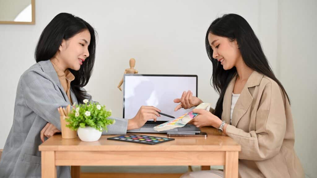Une jeune graphiste discutant avec une cliente en tenant dans sa main une palette de couleur. Elles sont devant un bureau sur lequel sont posés un ordinateur portable, une vase de fleur, un smartphone et un pot de crayon en bois.