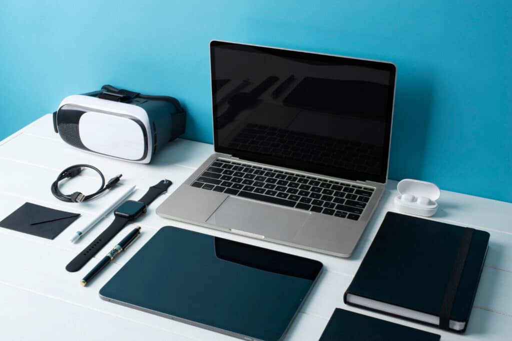 un ordinateur, des lunettes de réalité virtuelle, des écouteurs sans fil, une montre, un câble usb, un stylo, un crayon, un notebook et une tablette posés sur une table blanche.